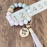Purple, Marble and grey silicone beaded wristlet with matching lobster clasp leather tassel on a ruler to show 3 inch width. Attached a large stainless steel circle charm tag engraved with Bride to Be, a small heart silhouette and the wedding date 4.18.24