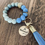 Blue, Marble and grey silicone beaded wristlet with matching lobster clasp leather tassel. Attached a large stainless steel circle charm tag engraved with Jr Bridesmaid, a small heart silhouette and the wedding date 4.18.24