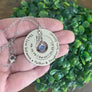 Silver stainless steel 1.5 inch round disc engraved with the memorial quote "The love between a daughter & father lasts forever" the inner circle at 1 inch and is engraved with date of birth 9.10.64 a heart symbol, Dad, heart symbol and the date of death 3.24.21. A September birthstone hangs in the center and an anel wing dangles from the top. Pendants hang from a stainless steel cable chain