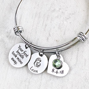 Silver triple loop wire bracelet with a round 3/4 inch engraved with "your first breath took mine away", a 5/8 inch round disc with engraved name "leon" and an image of a mom holding a baby, a 3/4 inch heart engraved with the birthday date "8.4.18", and a august birthstone.