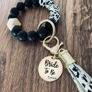 Cow pattern silicone beaded wristlet with matching lobster clasp leather tassel. Attached a large stainless steel circle charm tag engraved with Bride to Be, a engagement ring image and the wedding date 4.18.24