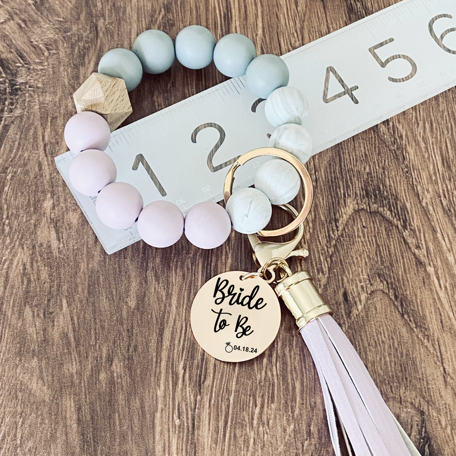 Purple, Marble and grey silicone beaded wristlet with matching lobster clasp leather tassel on a ruler to show 3 inch width. Attached a large stainless steel circle charm tag engraved with Bride to Be, a small heart silhouette  and the wedding date 4.18.24