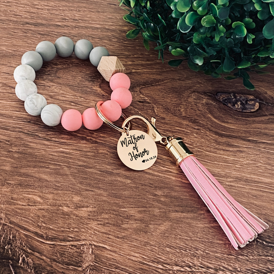 Pink, Marble and grey silicone beaded wristlet with matching lobster clasp leather tassel. Attached a large stainless steel circle charm tag engraved with Matron of Honor, a small heart silhouette  and the wedding date 4.18.24
