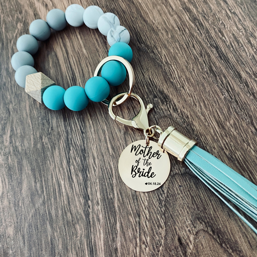 Teal, Marble and grey silicone beaded wristlet with matching lobster clasp leather tassel. Attached a large stainless steel circle charm tag engraved with Mother of the Bride, a small heart silhouette  and the wedding date 4.18.24