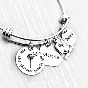 Silver Stainless Steel Engraved charm bracelet. circle disc engraved with "Oh, the places you'll go..." with a dandelion image. Next is a heart name charm. next is a graduation cap and tassle charm. lastly is a circle charm with the year 2024 engraved. All charms are attached to a triple loop stainless steel bangle bracelet