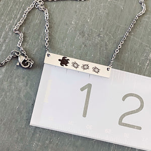 View of a Silver horizontal bar necklace engraved with one mom turtle and 3 baby turtles. The bar is attached to a silver stainless steel cable chain on top of a ruler to show the length of the bar at 1 3/8 inch 