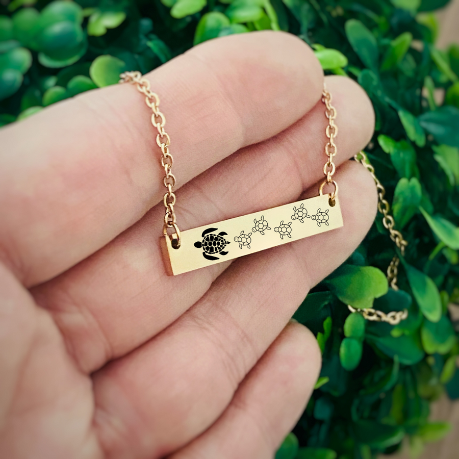 top side view of a yellow gold horizontal bar necklace engraved with one mom turtle and 5 baby turtles. The bar is attached to a yellow gold stainless steel cable chain