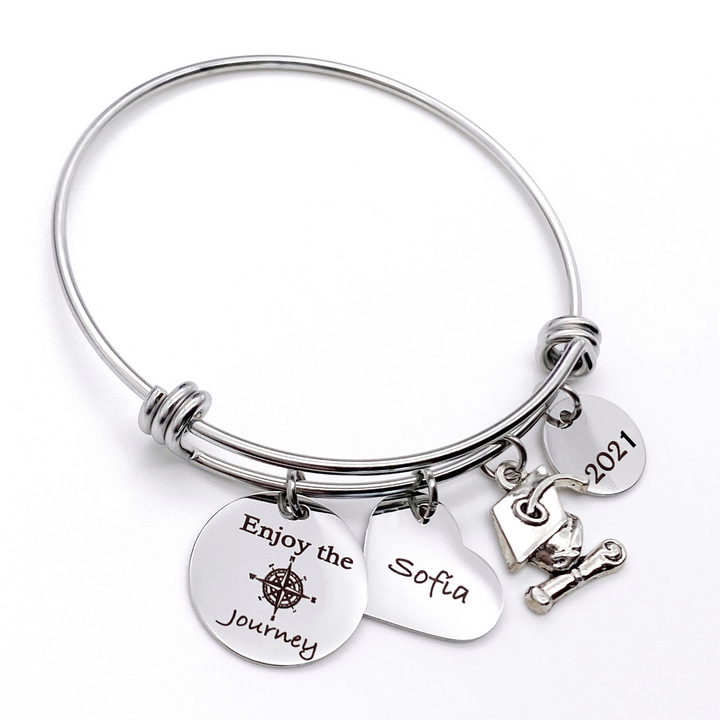 Silver Stainless Steel Engraved charm bracelet. circle disc engraved with "Enjoy the Journey" with a compass image. Next is a heart name charm. next is a graduation cap and tassel charm. lastly is a circle charm with the year "2021" engraved. All charms are attached to a triple loop stainless steel bangle bracelet