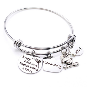 Silver Stainless Steel Engraved charm bracelet. circle disc engraved with "Every dream begins with a wish" with a dandelion image. Next is a heart name charm. next is a graduation cap and tassel charm. lastly is a circle charm with the year "2021" engraved. All charms are attached to a triple loop stainless steel bangle bracelet