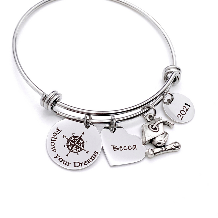 Silver Stainless Steel Engraved charm bracelet. circle disc engraved with "Follow your Dreams" with a compass image. Next is a heart name charm. next is a graduation cap and tassel charm. lastly is a circle charm with the year "2021" engraved. All charms are attached to a triple loop stainless steel bangle bracelet
