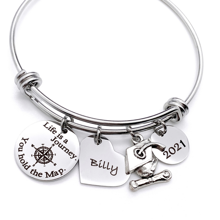 Silver Stainless Steel Engraved charm bracelet. circle disc engraved with "life is a journey. you hold the map" with a compass image. Next is a heart name charm. next is a graduation cap and tassle charm. lastly is a circle charm with the year "2021" engraved. All charms are attached to a triple loop stainless steel bangle bracelet