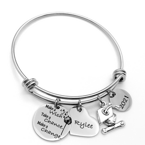 Silver Stainless Steel Engraved charm bracelet. circle disc engraved with "Make a wish. Take a chance. make a change" with a dandelion image. Next is a heart name charm. next is a graduation cap and tassel charm. lastly is a circle charm with the year "2021" engraved. All charms are attached to a triple loop stainless steel bangle bracelet