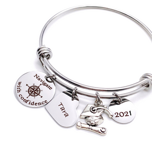 Silver Stainless Steel Engraved charm bracelet. circle disc engraved with "Navigate with Confidence" with a compass image. Next is a heart name charm. next is a graduation cap and tassel charm. lastly is a circle charm with the year "2021" engraved. All charms are attached to a triple loop stainless steel bangle bracelet