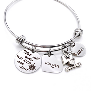 Silver Stainless Steel Engraved charm bracelet. circle disc engraved with "Not all who wander are lost" with a compass image. Next is a heart name charm. next is a graduation cap and tassel charm. lastly is a circle charm with the year "2021" engraved. All charms are attached to a triple loop stainless steel bangle bracelet