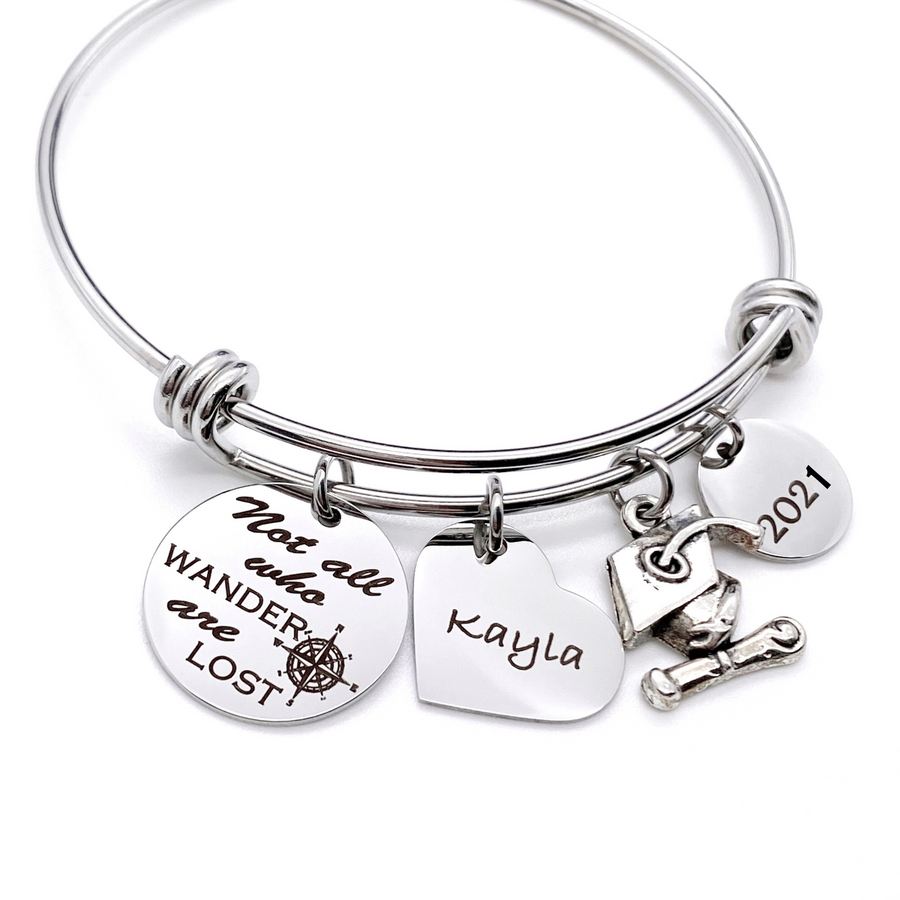 Silver Stainless Steel Engraved charm bracelet. circle disc engraved with "Not all who wander are lost" with a compass image. Next is a heart name charm. next is a graduation cap and tassel charm. lastly is a circle charm with the year "2021" engraved. All charms are attached to a triple loop stainless steel bangle bracelet