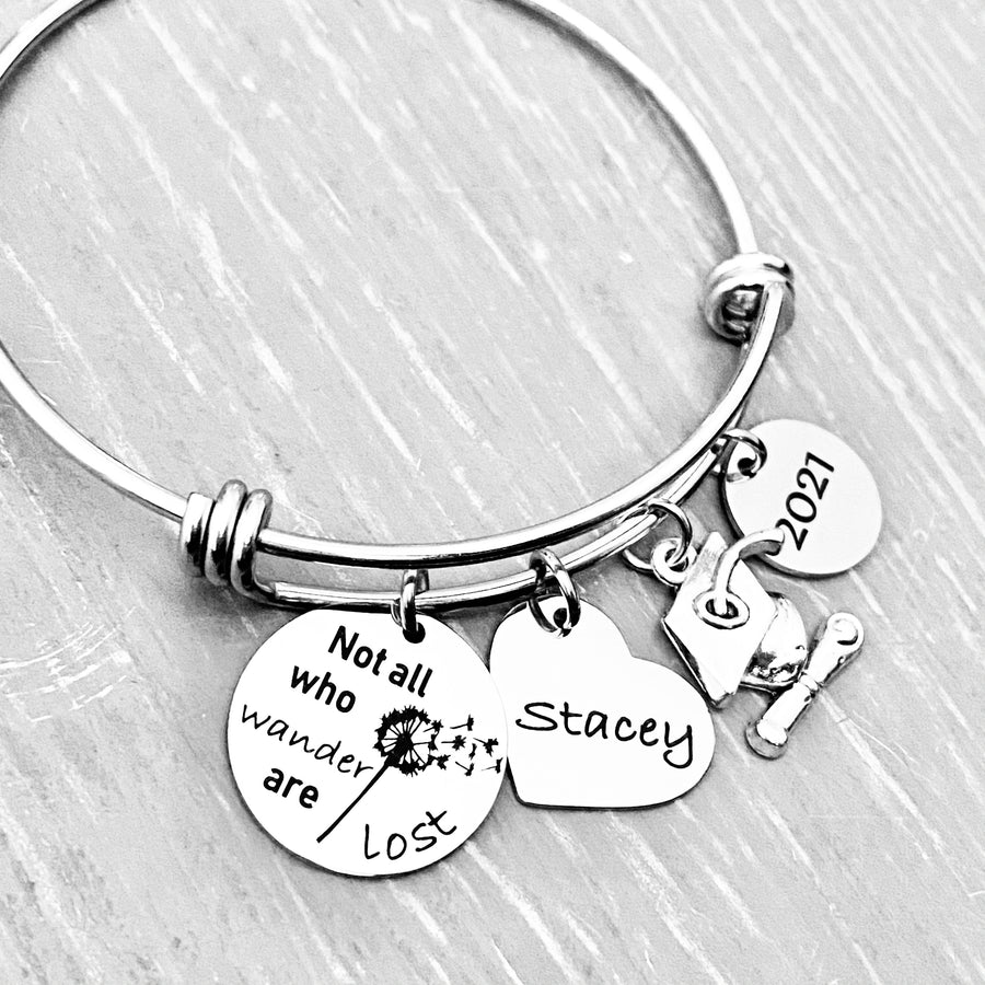 Silver Stainless Steel Engraved charm bracelet. circle disc engraved with "Not all who wander are lost" with a dandelion image. Next is a heart name charm. next is a graduation cap and tassel charm. lastly is a circle charm with the year "2021" engraved. All charms are attached to a triple loop stainless steel bangle bracelet