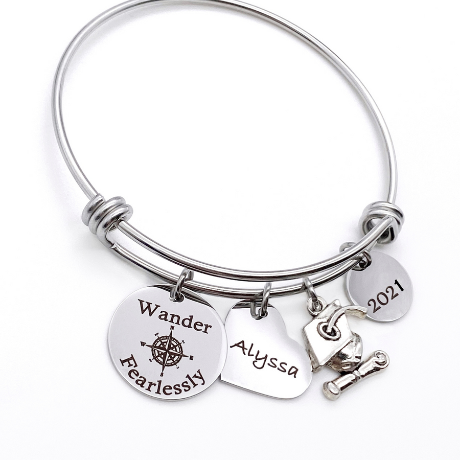 Silver Stainless Steel Engraved charm bracelet. circle disc engraved with "Wander Fearlessly" with a compass image. Next is a heart name charm. next is a graduation cap and tassel charm. lastly is a circle charm with the year "2021" engraved. All charms are attached to a triple loop stainless steel bangle bracelet