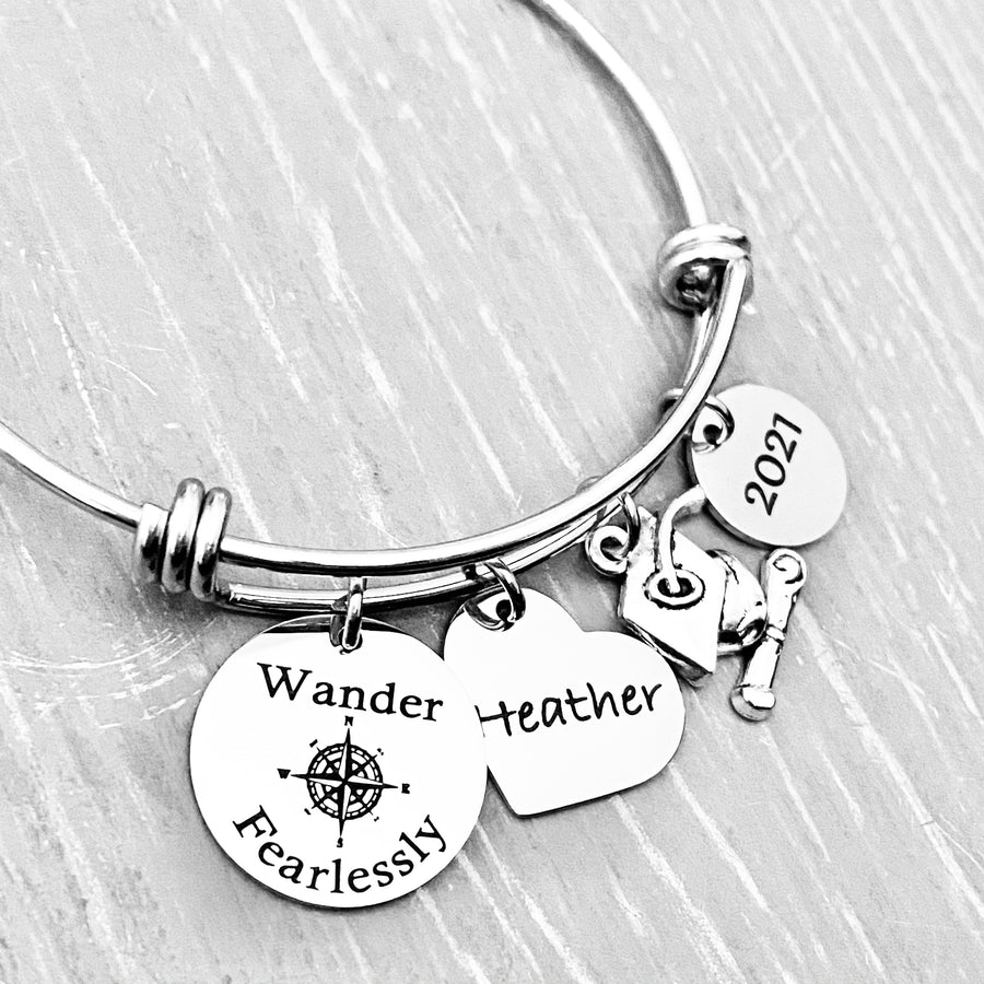Silver Stainless Steel Engraved charm bracelet. circle disc engraved with "Wander Fearlessly" with a compass image. Next is a heart name charm. next is a graduation cap and tassel charm. lastly is a circle charm with the year "2021" engraved. All charms are attached to a triple loop stainless steel bangle bracelet