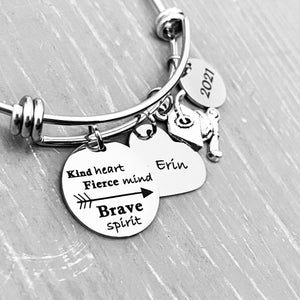Silver Stainless Steel Engraved charm bracelet. circle disc engraved with "Kind Heart. Fierce Mind. Brave Spirit" with a arrow image. Next is a heart name charm. next is a graduation cap and tassel charm. lastly is a circle charm with the year "2021" engraved. All charms are attached to a triple loop stainless steel bangle bracelet