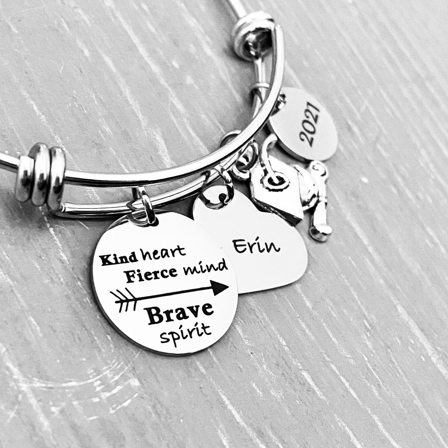 Silver Stainless Steel Engraved charm bracelet. circle disc engraved with "Kind Heart. Fierce Mind. Brave Spirit" with a arrow image. Next is a heart name charm. next is a graduation cap and tassel charm. lastly is a circle charm with the year "2021" engraved. All charms are attached to a triple loop stainless steel bangle bracelet