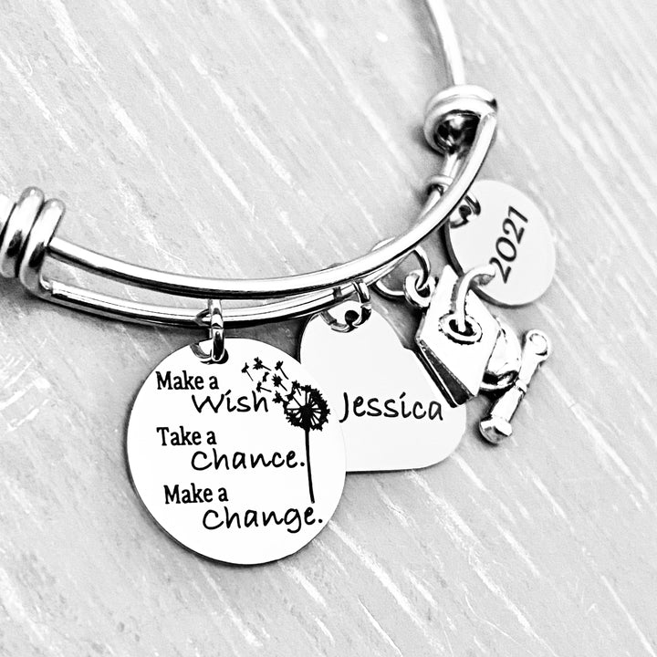 Silver Stainless Steel Engraved charm bracelet. circle disc engraved with "Make a wish. Take a chance. make a change" with a dandelion image. Next is a heart name charm. next is a graduation cap and tassel charm. lastly is a circle charm with the year "2021" engraved. All charms are attached to a triple loop stainless steel bangle bracelet