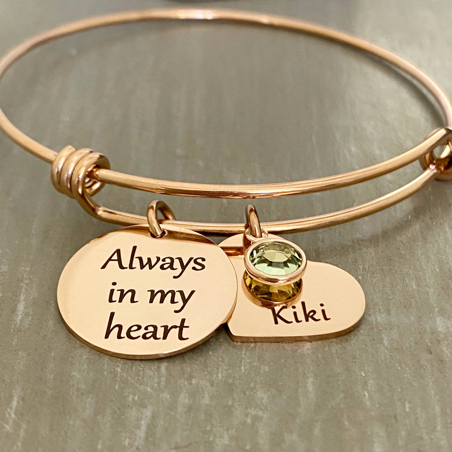 rose gold triple loop bangle charm bracelet. engraved on a 3/4" round charm is the verbiage "always in my heart". next to that is a 3/4" heart charm engraved with the name "kiki" and a august birthstone on top.