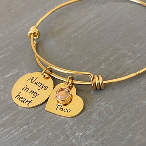 Yellow Gold stainless steel bangle charm bracelet with one round charm engraved with "always in my heart." next is a heart name charm engraved with "Theo" and a june birthstone