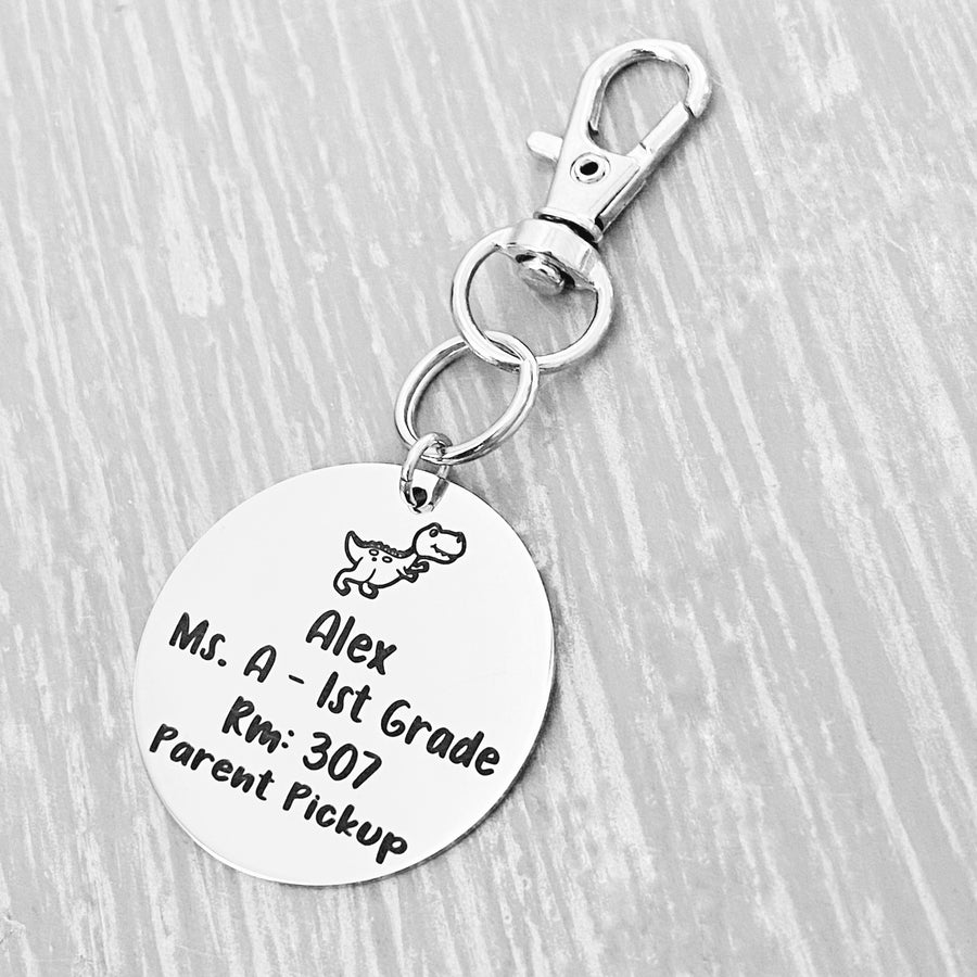 silver engraved stainless steel backpack tag keychain. engraved with a Dino Image, Name Alex. Teachers name Ms A 1st grade. Room number 307. and Parent Pickup