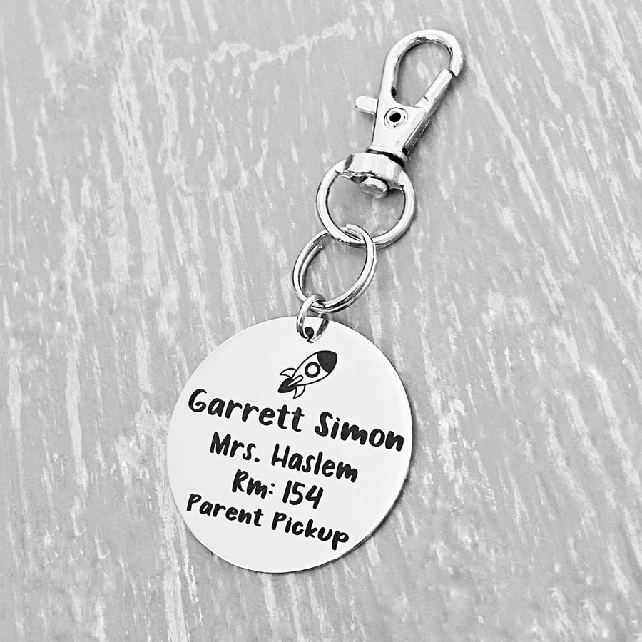 silver engraved stainless steel backpack tag keychain. engraved with a Rocket Image, Name Garrett Simon. Teachers name Mrs Haslem. Room number 154. and Parent Pickup