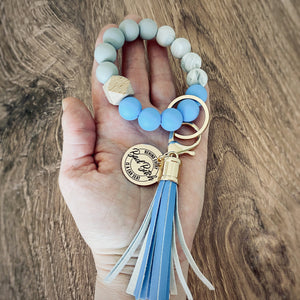 medium blue, white, and grey round silicone beaded wristlet with large lobster hook leather tassel that matches. A rose gold charm attached engraved with "Behind every bad bitch is a car seat" 
