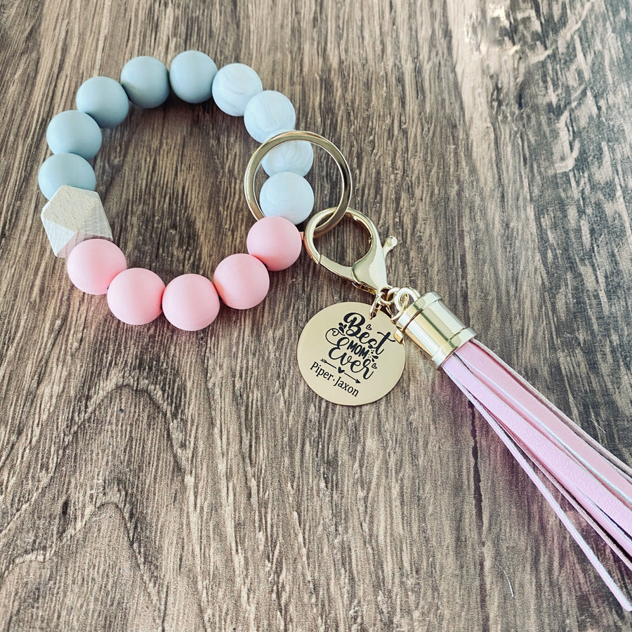 pink, white, and grey round silicone beaded wristlet with large lobster hook leather tassel that matches. A rose gold charm attached engraved with "Best Mom Ever" and personalized with the names "Piper and Jaxon""
