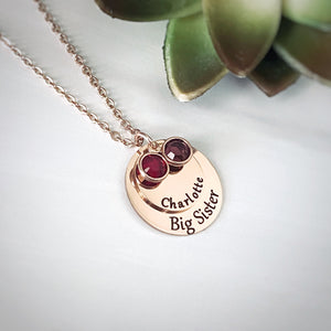 Rose Gold round pendant engraved with "Big Sister". A smaller round pendant lays on top with the name "Charlotte". On top of that pendant sits 2 birthstones. One July and one february. All charms are attached to a rose gold stainless steel cable chain