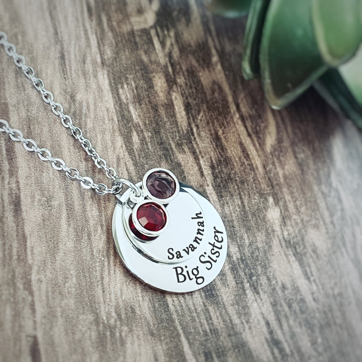 Silver round pendant engraved with "Big Sister". A smaller round pendant lays on top with th ename "Savannah". Ontop of that pendant sits 2 birthstones. One July and one February. All charms are attached to a silver stainless steel cable chain