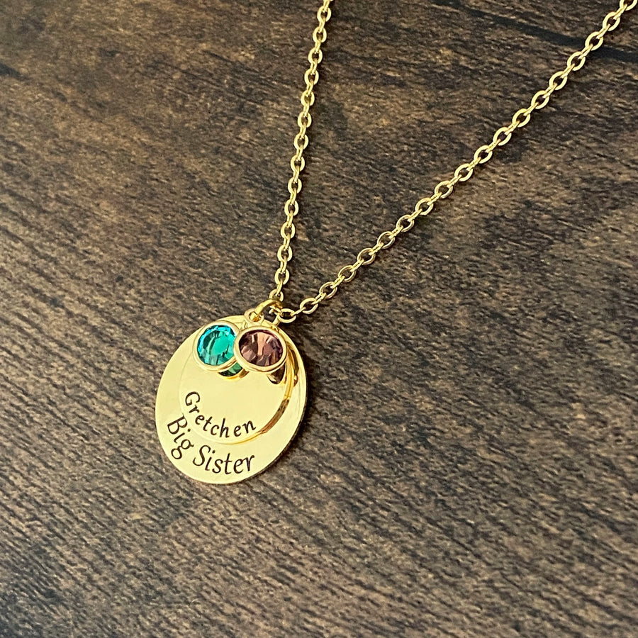 Yellow Gold round pendant engraved with "Big Sister". A smaller round pendant lays on top with the name "Gretchen". On top of that pendant sits 2 birthstones. One December and one February. All charms are attached to a yellow gold stainless steel cable chain
