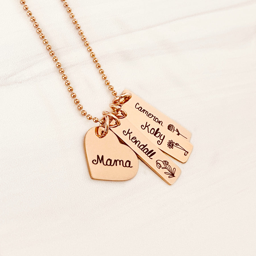 Rose Gold necklace with a heart charm engraved with "mama". 3 rectangle charm pendants engraved with the name "Kendall" and Snow Drop january birth month flower. 2nd tag engraved with Koby and July water lily flower. 3rd tag with "Cameron" and November Chrysanthemum flower