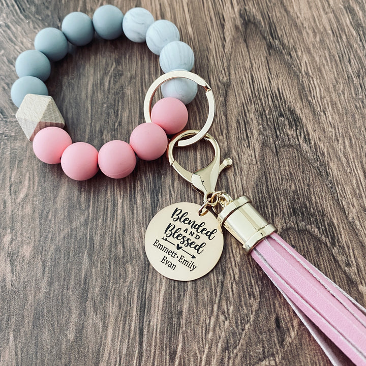 Pink, White, Grey round silicone beaded Wristlet with matching leather lobster clasp keychain hook. Attached is a rose gold charm tag engraved with "Blended and Blessed" with an arrow heart image and personalized with the names "Emmett, Emily, and Evan"