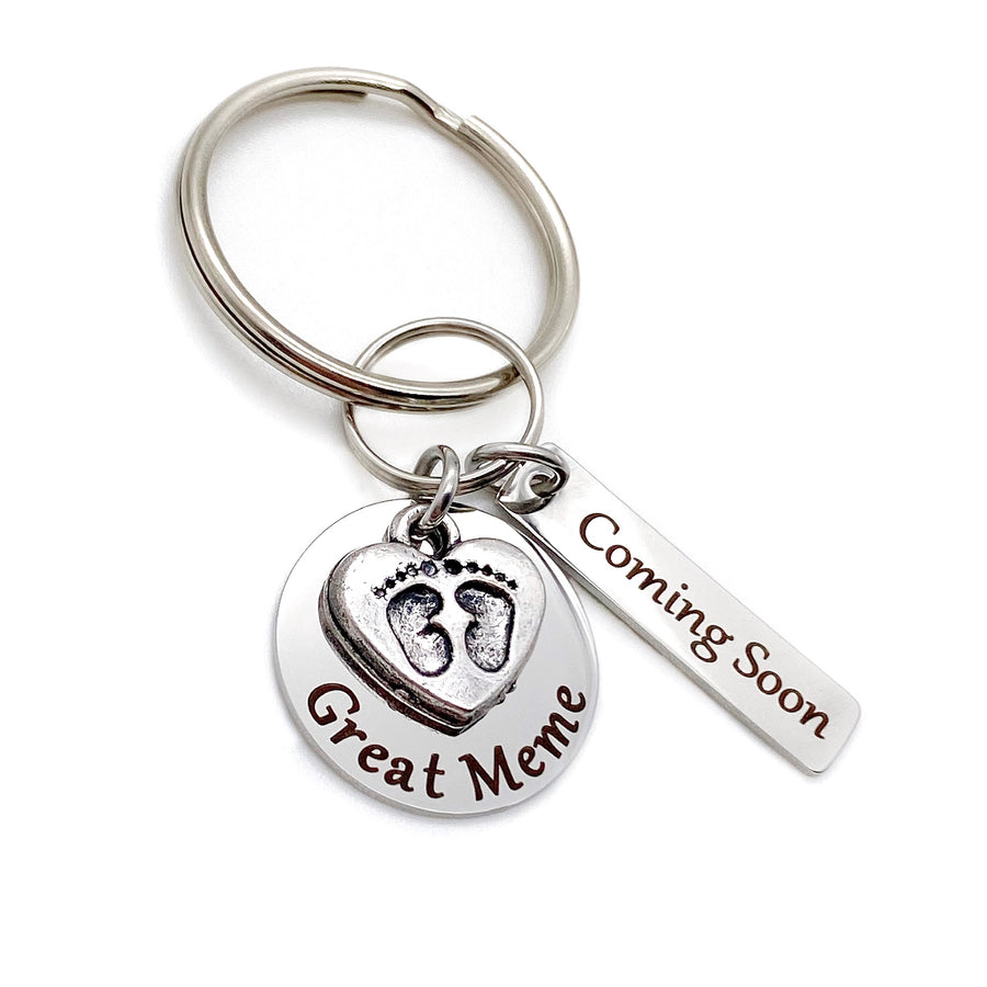 silver stainless steel keychain. circle disc engraved with great meme. baby's feet charm lays on top. a 1 inch rectangle tage engraved with Coming Soon.