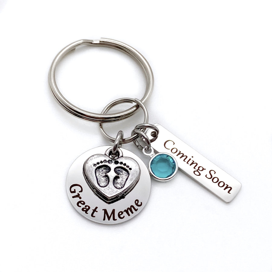 silver stainless steel keychain. circle disc engraved with great meme. baby's feet charm lays on top. besides is a december stone. a 1 inch rectangle tag engraved with Coming Soon.