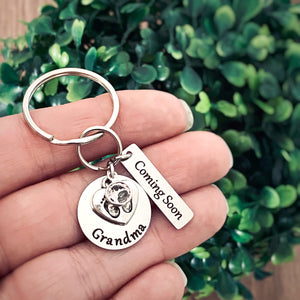 silver stainless steel keychain. circle disc engraved with "grandma". baby's feet charm lays on top. besides is a april stone. a 1 inch rectangle charm tag engraved with Coming Soon.