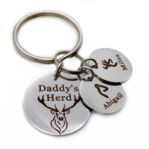 silver stainless steel 7/8" round keychain engraved with a Buck Deer image and "Dad's Herd". Next to the main charm are 2 small 1/2" name tags. first tag has a fawn deer head image and name Abigail. Second charm with a boy deer charm with name Miles.