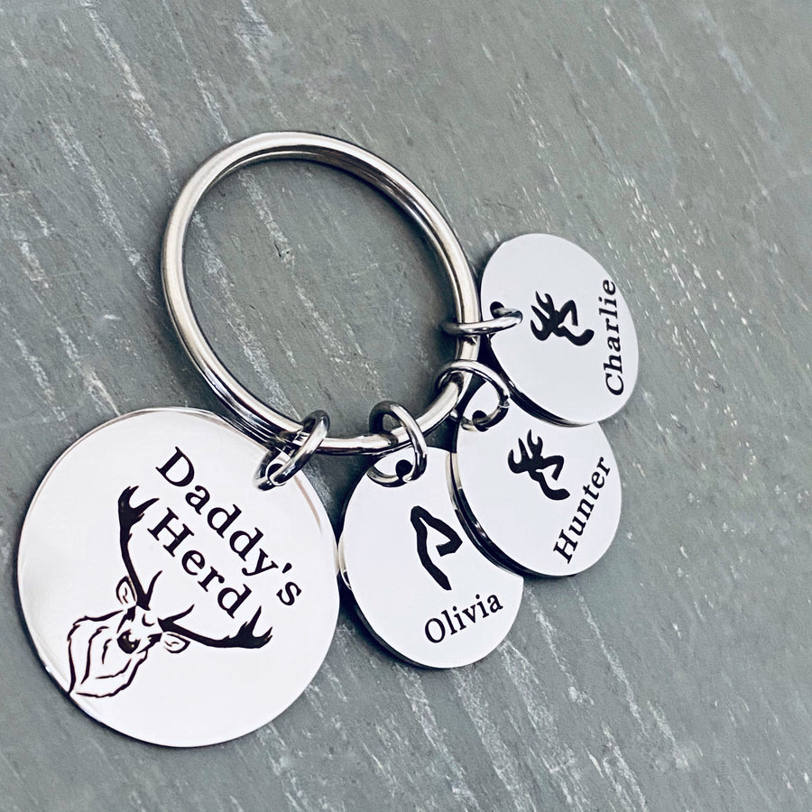 silver stainless steel 7/8" round keychain engraved with a Buck Deer image and "Daddy's Herd". Next to the main charm are 3 small 1/2" name tags. first tag has a fawn deer head image and name Olivia. Second charm with a male deer charm with name Hunter. third charm is a boy deer and the name Charlie.