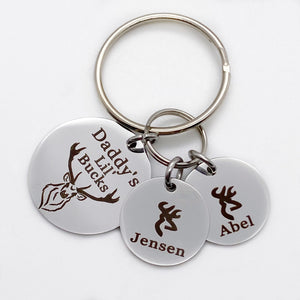 silver stainless steel 7/8" round keychain engraved with a Buck Deer image and "Daddy's Lil' Bucks". Next to the main charm are 3 small 1/2" name tags. first tag has a baby deer head image and name Jensen. Second charm with a male deer charm with name Abel.