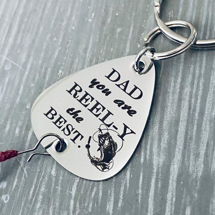 Close up of a 1 inch silver stainless steel fishing lure keychain. Engraved with DAD you are REEL-Y the BEST" attached to a durable steel keyring and fishing feather.