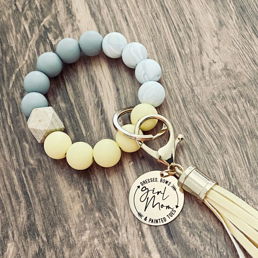 Yellow, white, grey silicone beaded bracelet with leather lobster hook keychain tassel. Engraved round charm tag with "Girl Mom. Dresses. Bows & painted Toes"
