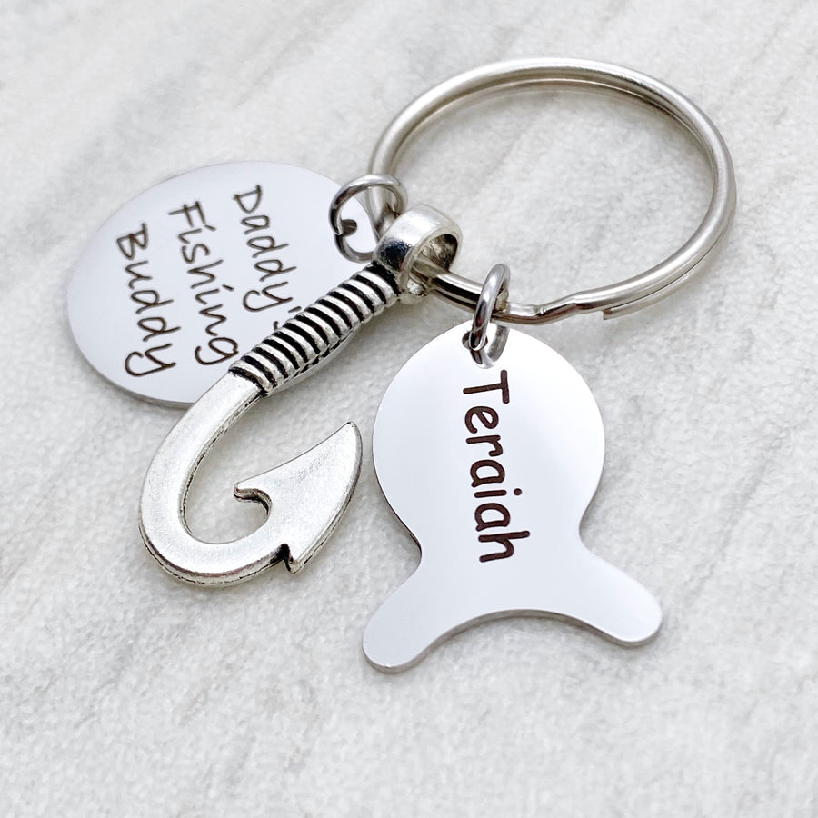 Silver engraved personalized fishing keychain. Main tag is engraved with "Daddy's fishing buddy". Next is a silver fishing hook. Next is a silver engraved fish tag charm with the name "Teraiah"