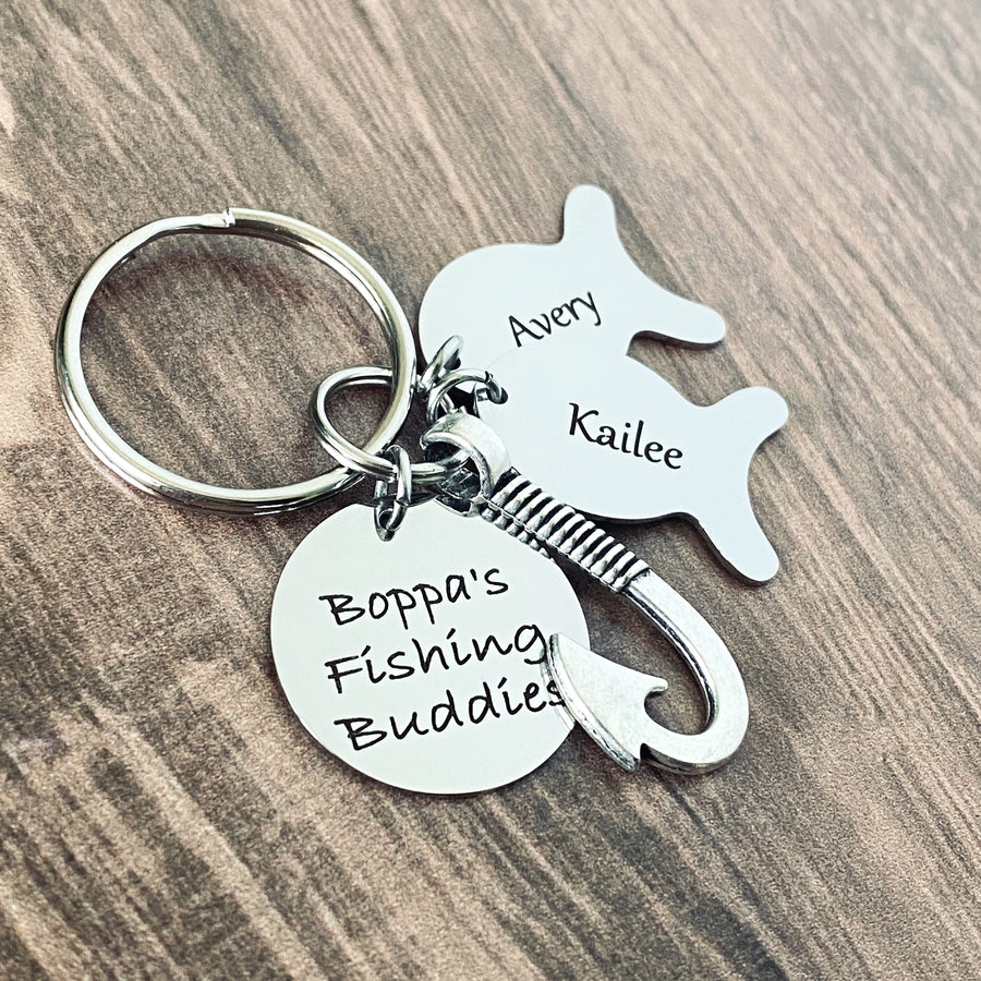 Silver engraved personalized fishing keychain. Main tag is engraved with "Boppa's fishing buddy". Next is a silver fishing hook. Next is a silver engraved fish tag charm with the name "Kailee" and another fish charm engraved with the name "Avery""