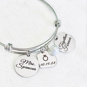 Silver stainless steel bangle charm bracelet with 3/4" laser engraved mrs. charm tag, a 3/4 inch heart tag with wedding ring and the wedding date 10.15.22, white pearl, and 3/4" engraved with "together forever"
