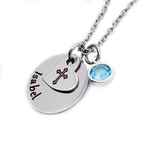 silver engraved 5/8" engraved round stainless steel disc with name isabel. ontop of the disc is a small 1/2" tilted heart with a religious engraved cross. both attached to a stainless steel silver cable chain. next to the pendant is a march swarovski birth stone