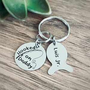round silver stainless steel disc engraved with "hooked on daddy" and a reel and rod fishing pole. Attached next is a fish style name tag engraved with the kids name Luis Jr all charms are added on 1 " stainless steel keyring