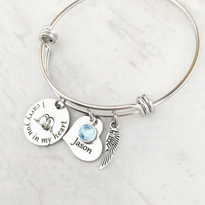 silver bangle charm bracelet with a 3/4" round engraved disc with the image of a baby sleeping under angel wings and the verbiage "I carry you in my heart" next to it is a 3/4" heart engraved with the name "Jason" and. a blue march birthstone. Last is an angel wing charm.